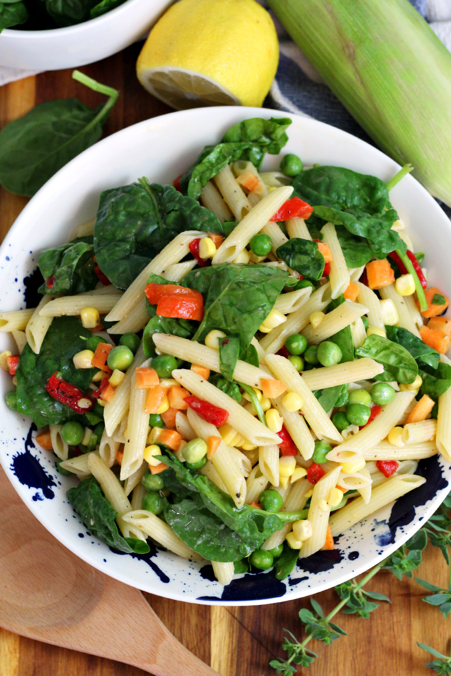 Top view of Vegetable Penne Pasta Salad in serving dish. Salad sits on wooden board. Wood spoon, baby spinach, lemon, ear of corn, and fresh oregano sprigs also in partial view.