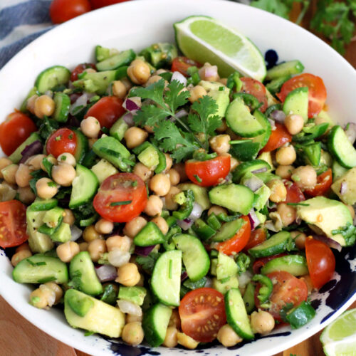 Cilantro Lime Chickpea Salad in serving dish sits on wooden board. Wooden serving spoon, fresh cilantro, lime wedges, and cherry tomatoes also in partial view.