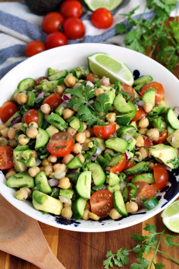 Cilantro Lime Chickpea Salad in serving dish sits on wooden board. Wooden serving spoon, fresh cilantro, lime wedges, and cherry tomatoes also in partial view.