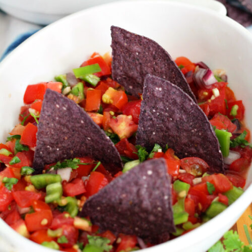 Shark Bite Chips and Fresh Salsa served in shallow dish. Blue corn chips stand upright in salsa. Blue tortilla chips in small bowl and fresh cilantro also in partial view.