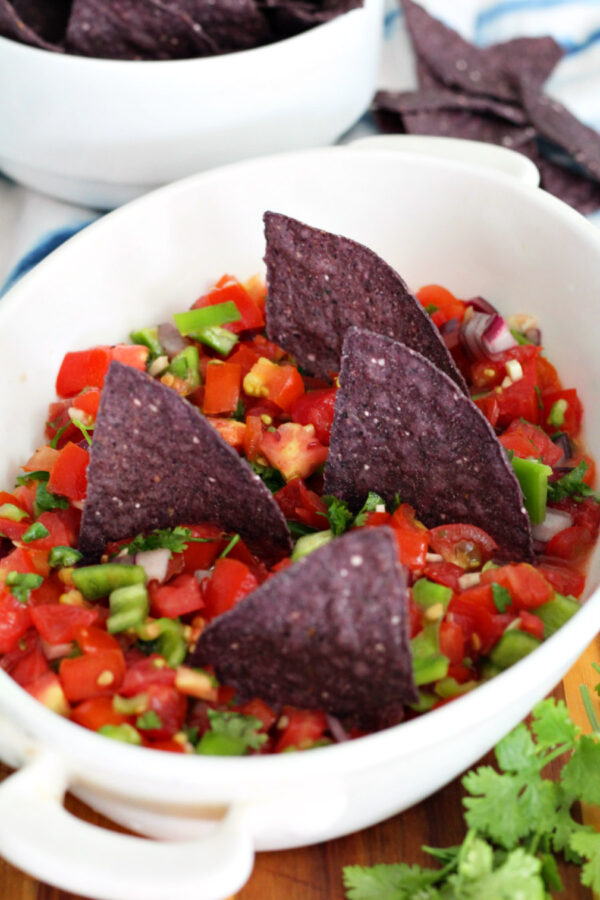 Shark Bite Chips and Fresh Salsa served in shallow dish. Blue corn chips stand upright in salsa. Blue tortilla chips in small bowl and fresh cilantro also in partial view.