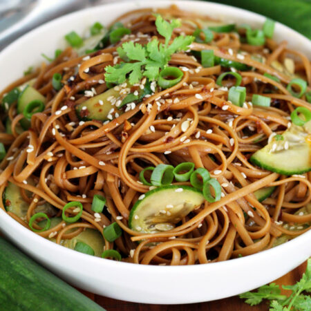 Sesame Cucumber Noodles sit in shallow serving dish on wooden board. Fresh cucumbers, metal tongs, and fresh cilantro also in partial view.