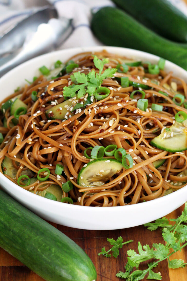 Sesame Cucumber Noodles sit in shallow serving dish on wooden board. Fresh cucumbers, metal tongs, and fresh cilantro also in partial view.