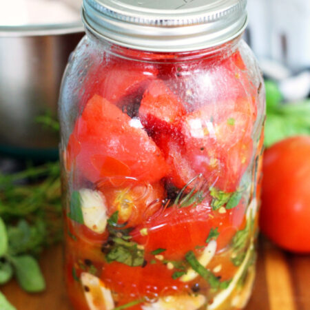 Italian Marinated Refrigerator Garden Tomatoes in quart mason jar. Jar sits on wooden board. Metal mixing bowl, tomatoes, and fresh herbs in partial view in background.