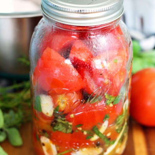 Italian Marinated Refrigerator Garden Tomatoes in quart mason jar. Jar sits on wooden board. Metal mixing bowl, tomatoes, and fresh herbs in partial view in background.