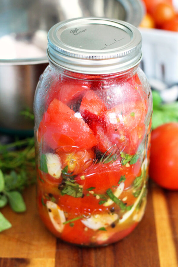 Italian Marinated Refrigerator Garden Tomatoes in quart mason jar. Jar sits on wooden board. Metal mixing bowl, tomatoes, and fresh herbs in partial view in background.