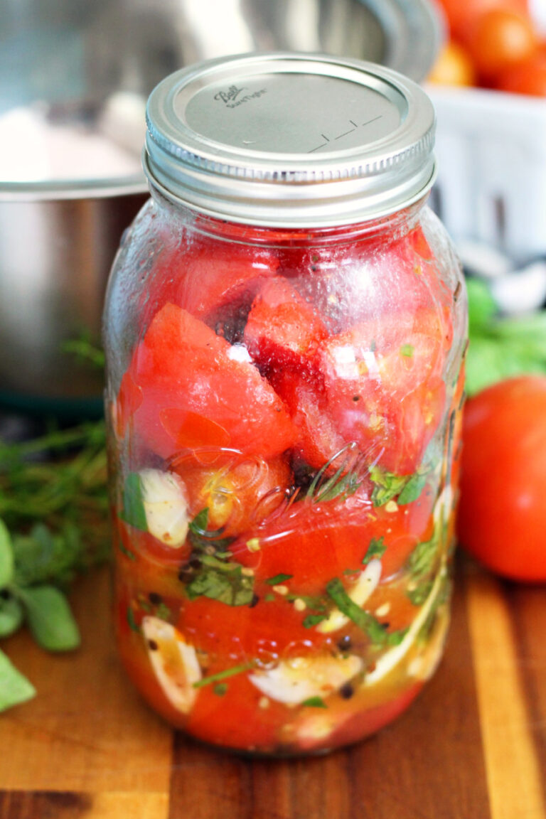 Italian Marinated Refrigerator Garden Tomatoes in quart mason jar. Jar sits on wooden board. Metal mixing bowl, tomatoes, and fresh herbs in partial view in background.
