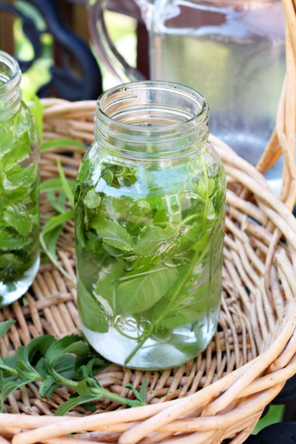 Mason jar filled with orange mint sun tea herbs and water sits inside basket. Partial view of another sun tea jar and glass pitcher in background.