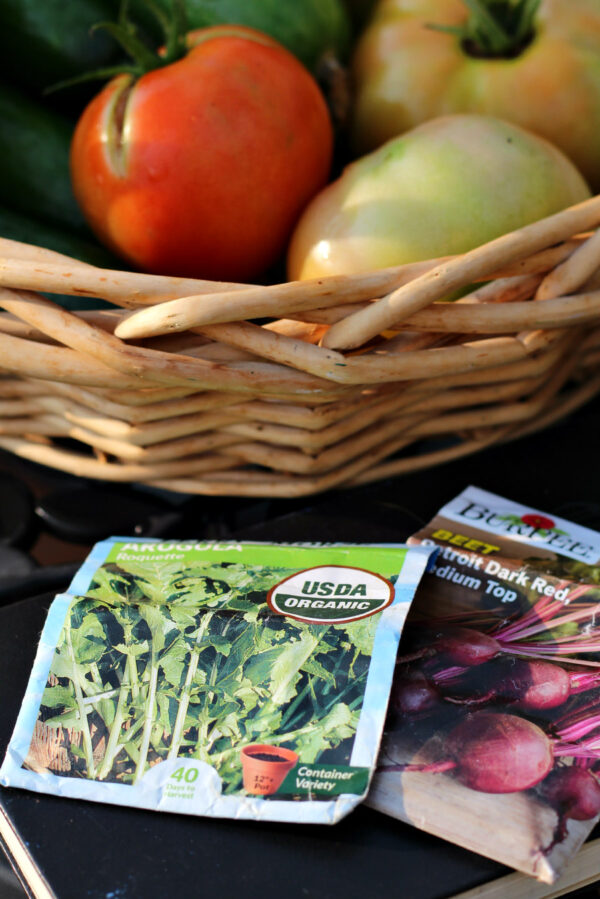 Arugula and beet seed packets sit on outdoor table in front of basket of fresh garden tomatoes and cucumbers.