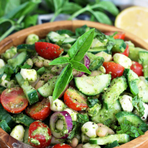 Italian Cucumber White Bean Salad with Pesto in wooden bowl on wooden board. Fresh basil leaves, cherry tomatoes, and lemon half in partial view in background.