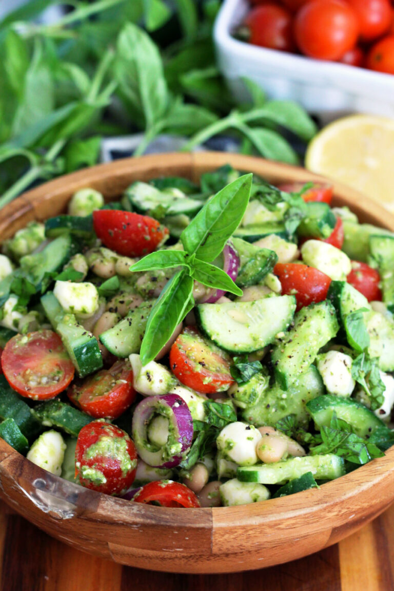 Italian Cucumber White Bean Salad with Pesto in wooden bowl on wooden board. Fresh basil leaves, cherry tomatoes, and lemon half in partial view in background.