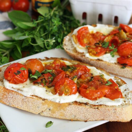 Burst Cherry Tomato and Herb Whipped Ricotta Toast on white serving dish on wooden board. Fresh herbs, hot honey bottle, and cherry tomatoes also in partial view.