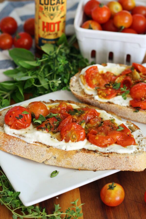 Burst Cherry Tomato and Herb Whipped Ricotta Toast on white serving dish on wooden board. Fresh herbs, hot honey bottle, and cherry tomatoes also in partial view.