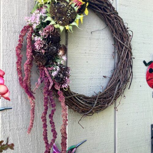 Angled view of Dried Flower Head Wreath on shed. Pink begonias, hummingbird, and ladybug decoration also in partial view.