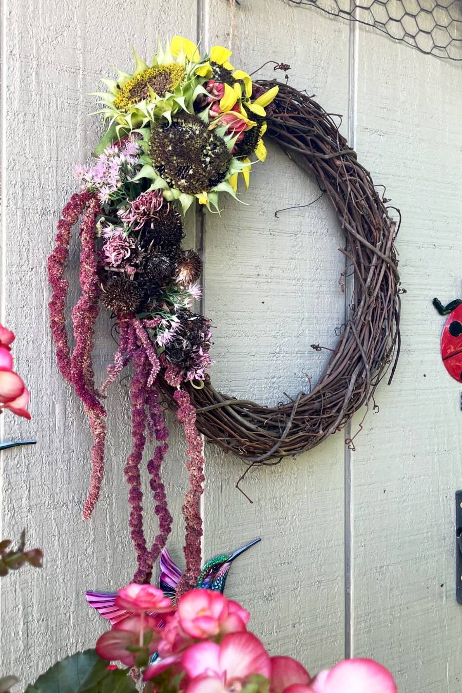 Angled view of Dried Flower Head Wreath on shed. Pink begonias, hummingbird, and ladybug decoration also in partial view.