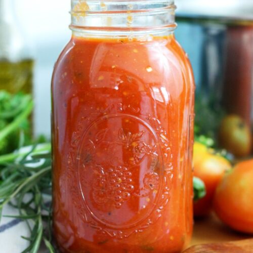 Large mason jar of homemade roasted garden tomato sauce sits on wooden board. Wooden spatula spoon, fresh herbs, and tomatoes also sit on board. Large stainless steel pot and olive oil bottle in partial view in background.