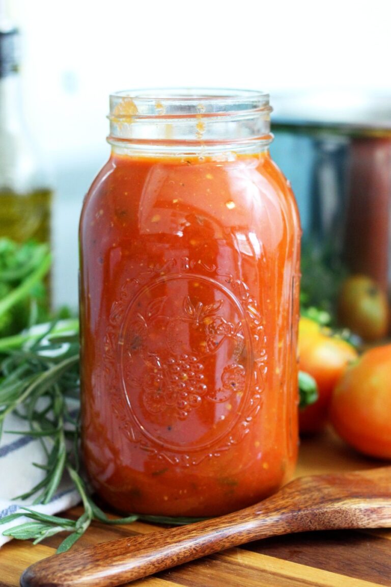 Large mason jar of homemade roasted garden tomato sauce sits on wooden board. Wooden spatula spoon, fresh herbs, and tomatoes also sit on board. Large stainless steel pot and olive oil bottle in partial view in background.